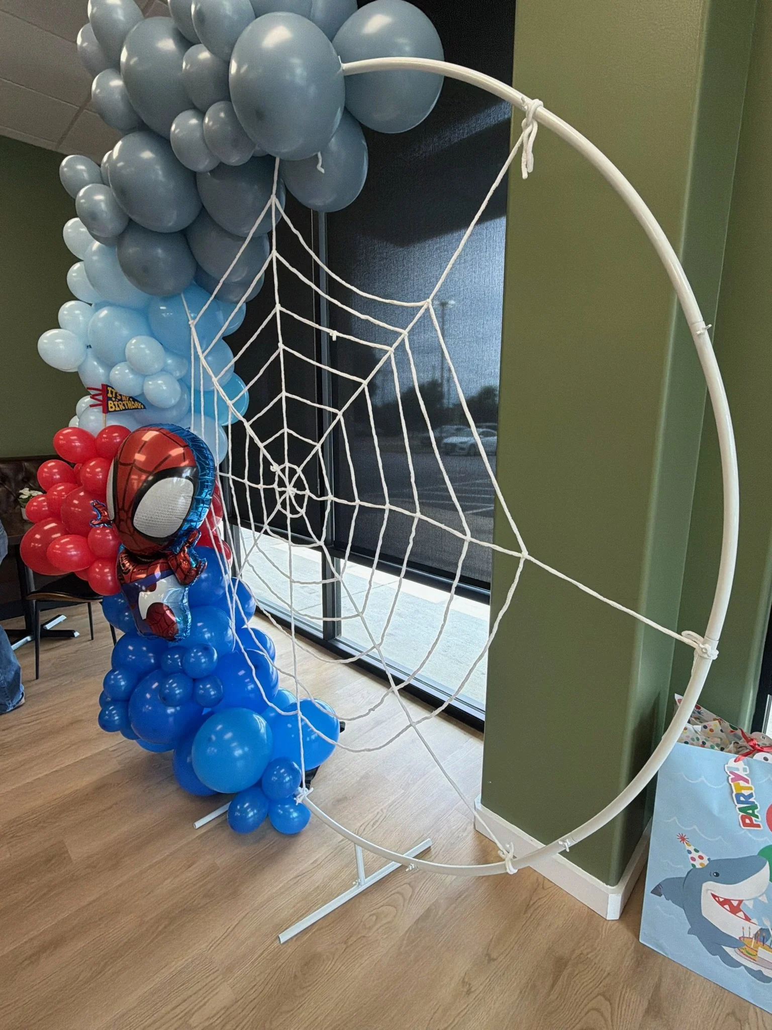 A balloon display with blue, gray, and red balloons, featuring a Spider-Man shaped balloon, next to a white web decoration, situated indoors near a window with blinds and a green wall.
