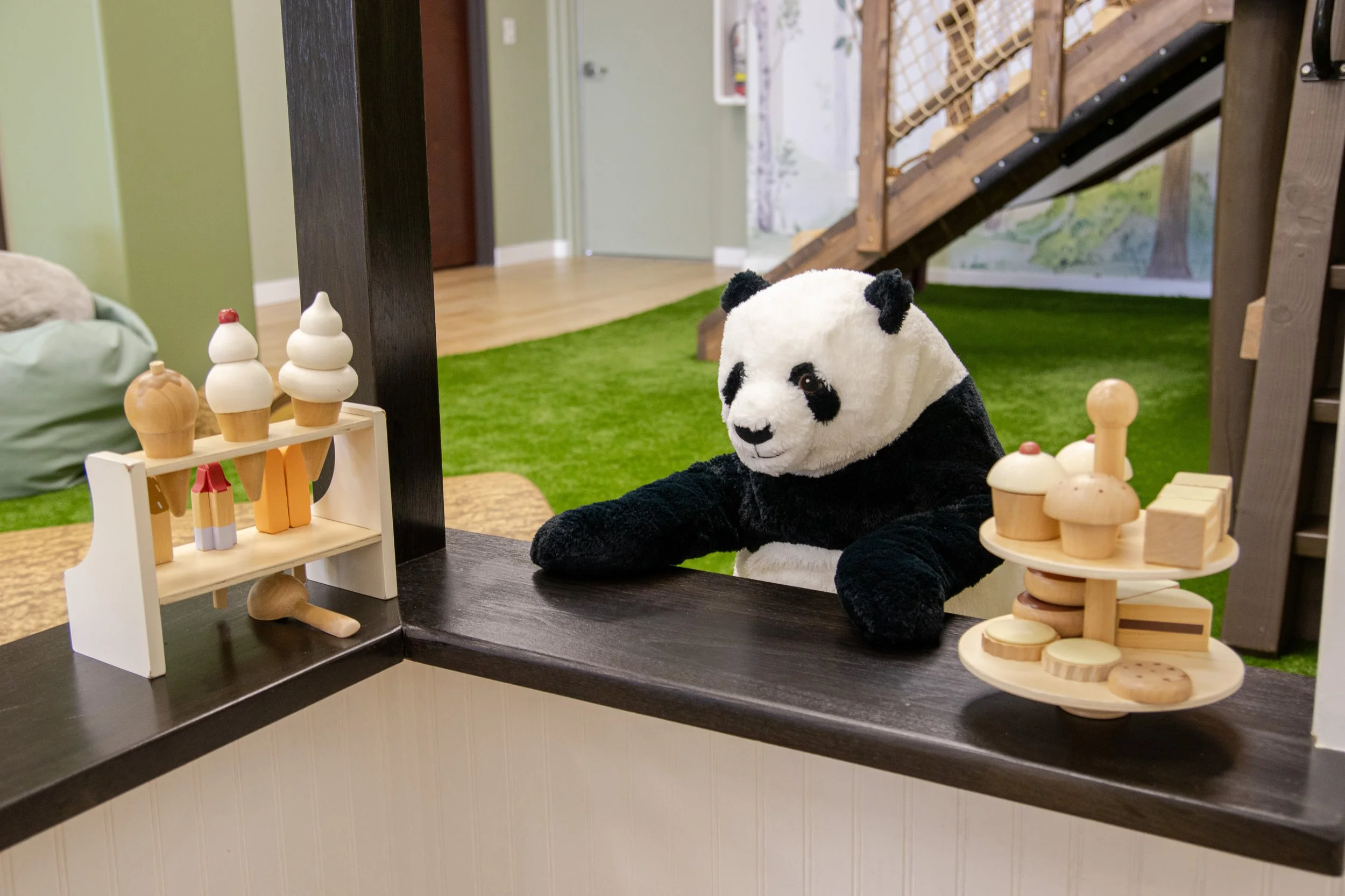 A stuffed panda bear sitting at a play kitchen counter with wooden toy food items, in a playroom with green carpet and wooden staircase.