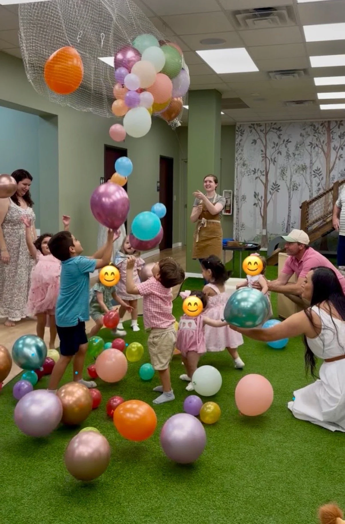 Children and adults at a birthday party, playing with colorful balloons in an indoor setting with a green carpeted floor, a wall with tree mural, and a ceiling with lights.