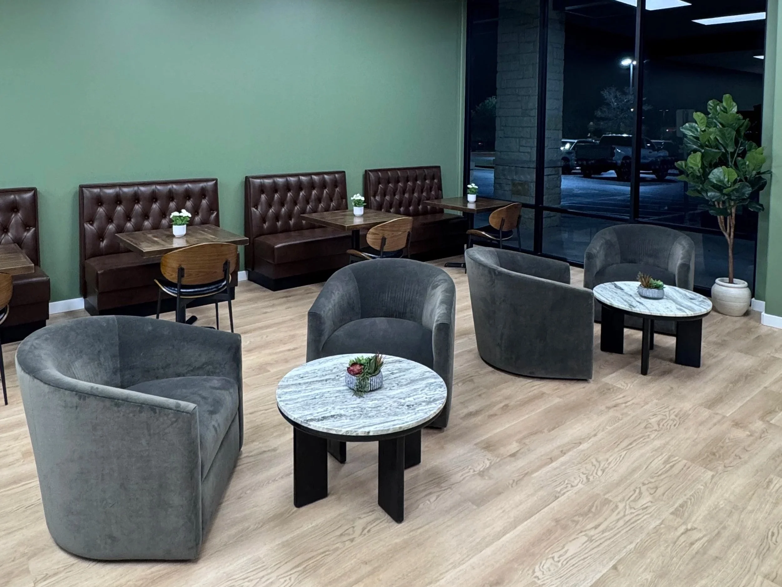 A cozy indoor seating area with gray velvet armchairs, small round marble-top tables with black bases, and potted plants on the tables. A green accent wall and large glass windows that reveal parked cars outside.