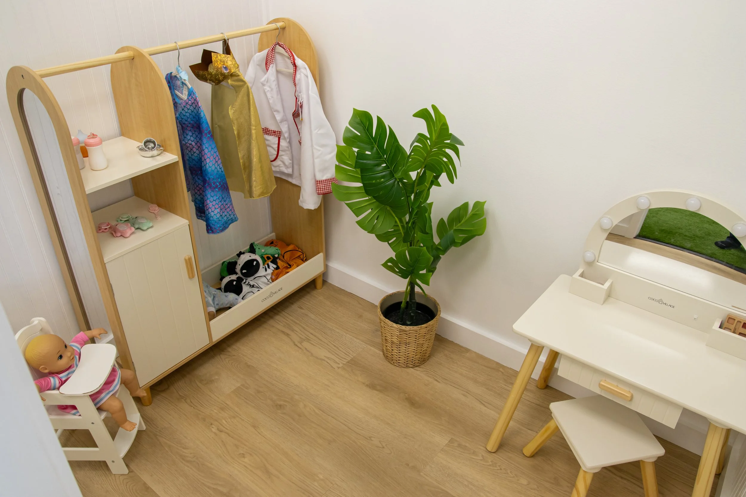 A children's playroom with a doll sitting in a high chair, a wooden shelf with toys and stuffed animals, a large potted Monstera plant, a white vanity with a mirror, and hanging costumes on a wooden rack.