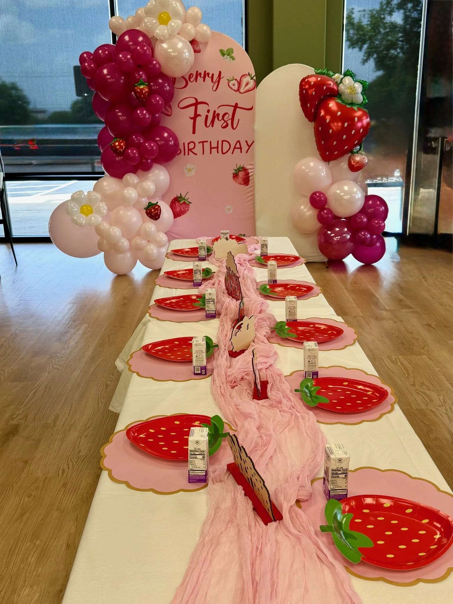Decorative table setup for a first birthday celebration with pink and red strawberry-themed plates, pink table runner, balloon arches with strawberries, white flowers, and a large pink sign reading 'First Birthday' with strawberry and flower illustra