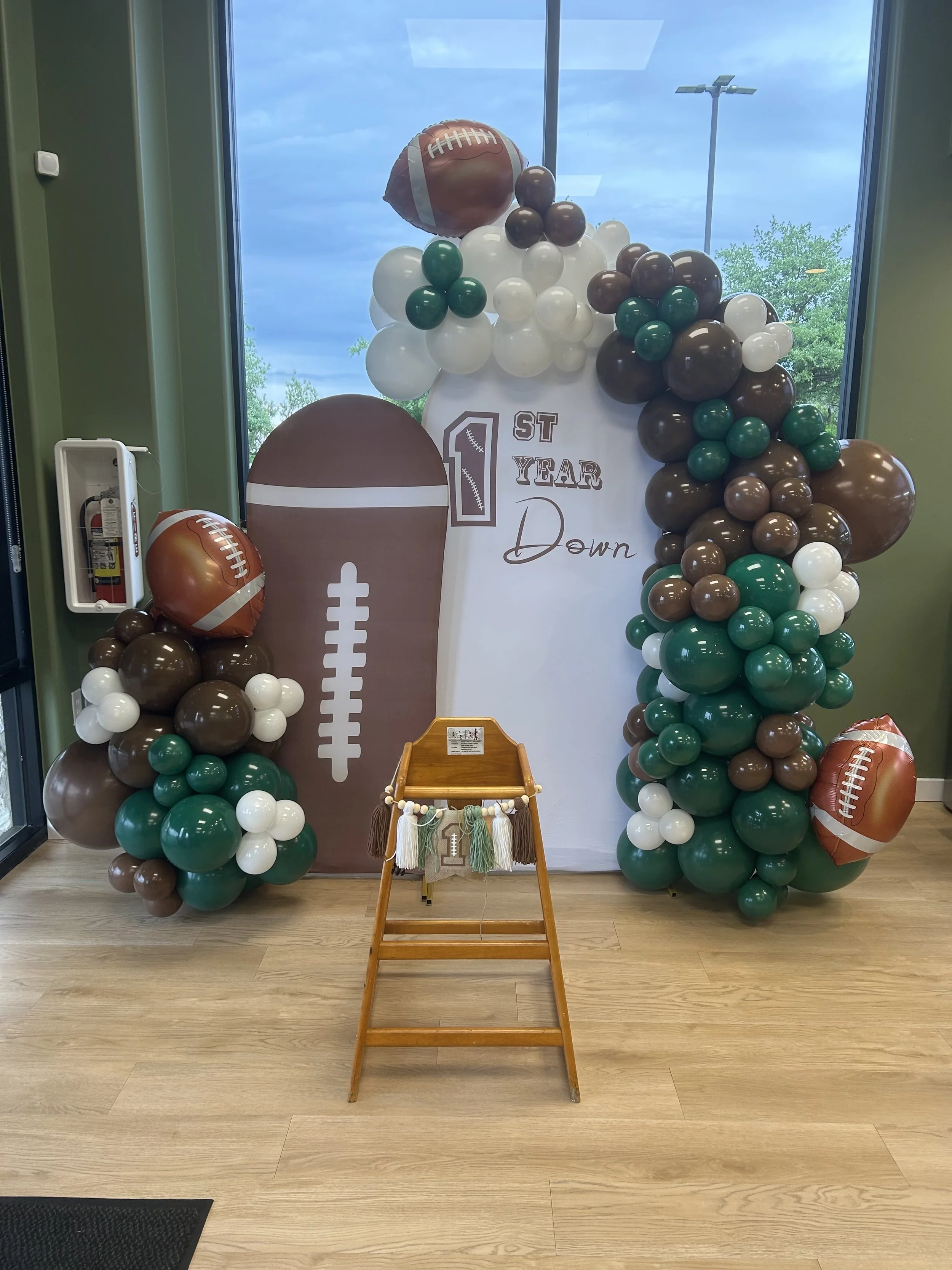 Decorative display for a first-year celebration featuring a balloon arch with football-themed balloons, a large football cutout, and a white backdrop with '1st Year Down' written on it.
