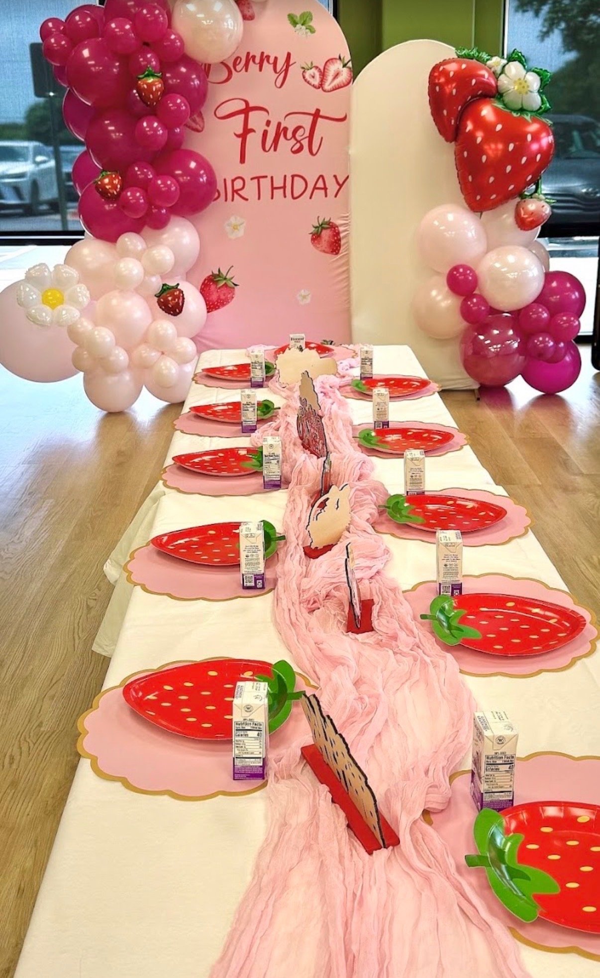 Birthday party decorations with a pink and white balloon arch, a banner with strawberries and the words 'Happy First Birthday,' and a table set with strawberry-themed plates, pink table runner, and strawberry juice cartons.