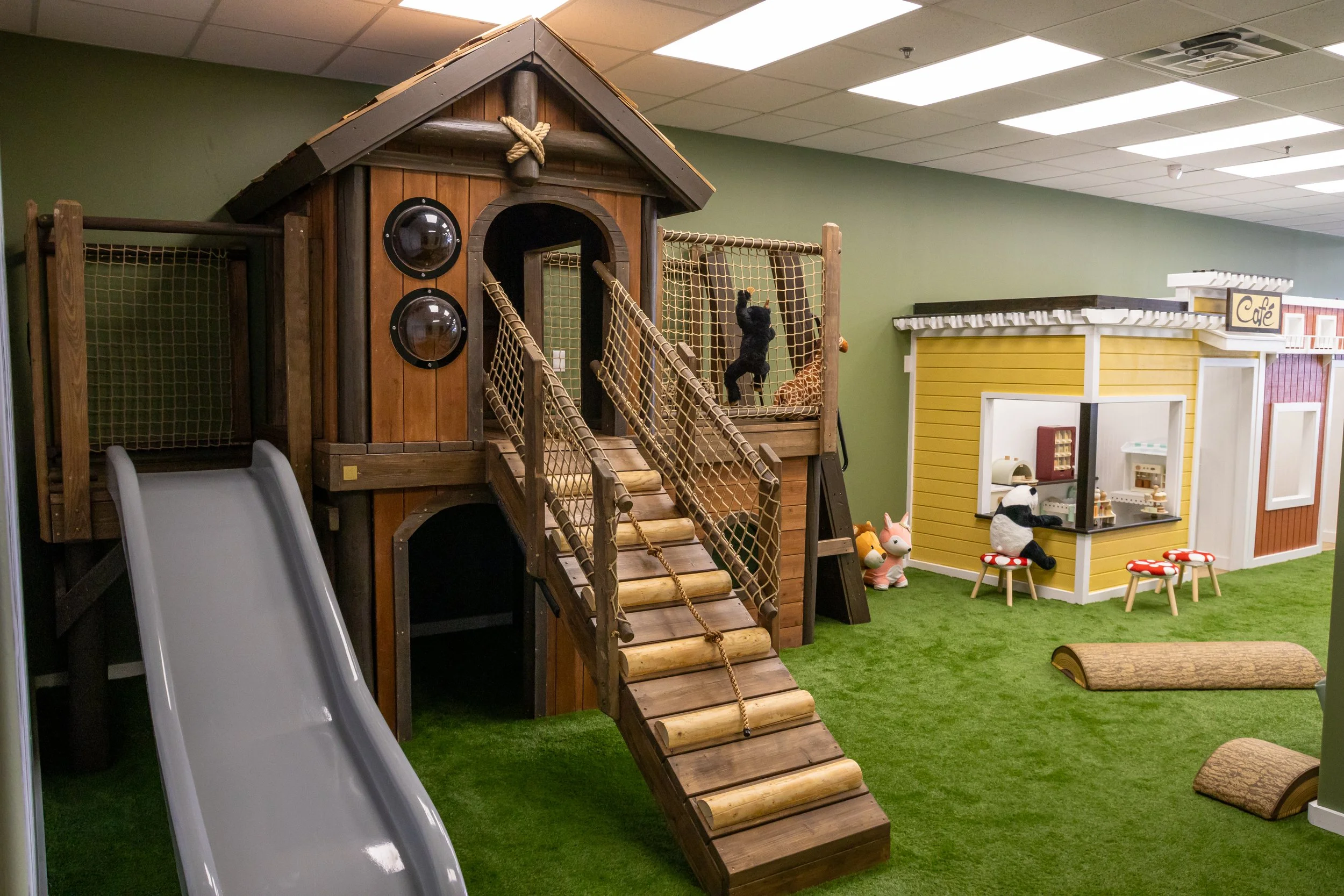 Indoor children's play area featuring a wooden playhouse with a slide, climbing ramp, and netting, with a green wall and various animal and café-themed decorations.