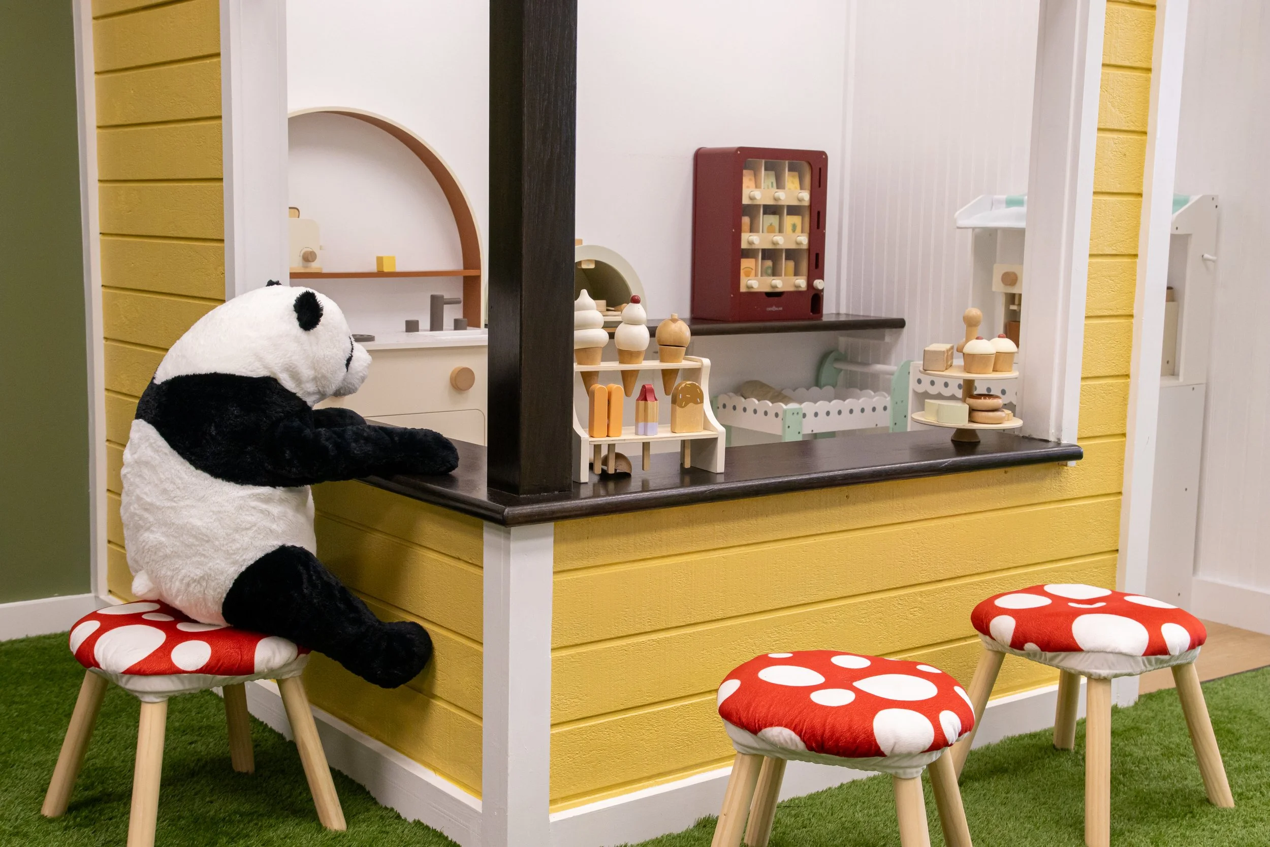 A small play kitchen area with wooden stools and a stuffed panda bear sitting on one of the stools. The play kitchen features shelves with toy food, a red toy oven, and a white sink, surrounded by yellow siding and white walls.