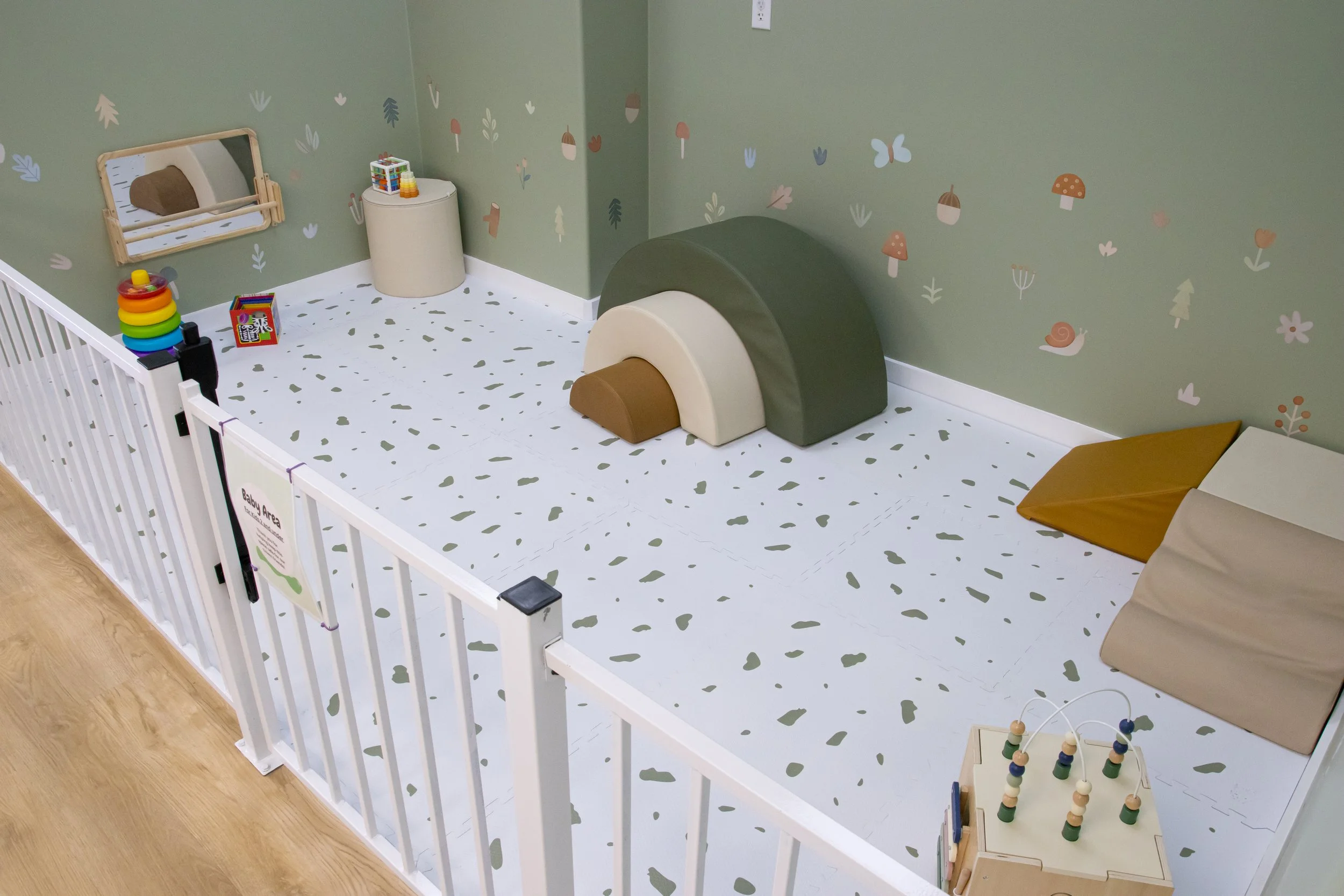 A children's play area with soft padded flooring, surrounded by a white safety fence. There are various toys including stacking rings, a small mirror, and a bead maze. The walls are painted green with playful nature-themed decals of mushrooms, leaves, butterflies, and a snail.