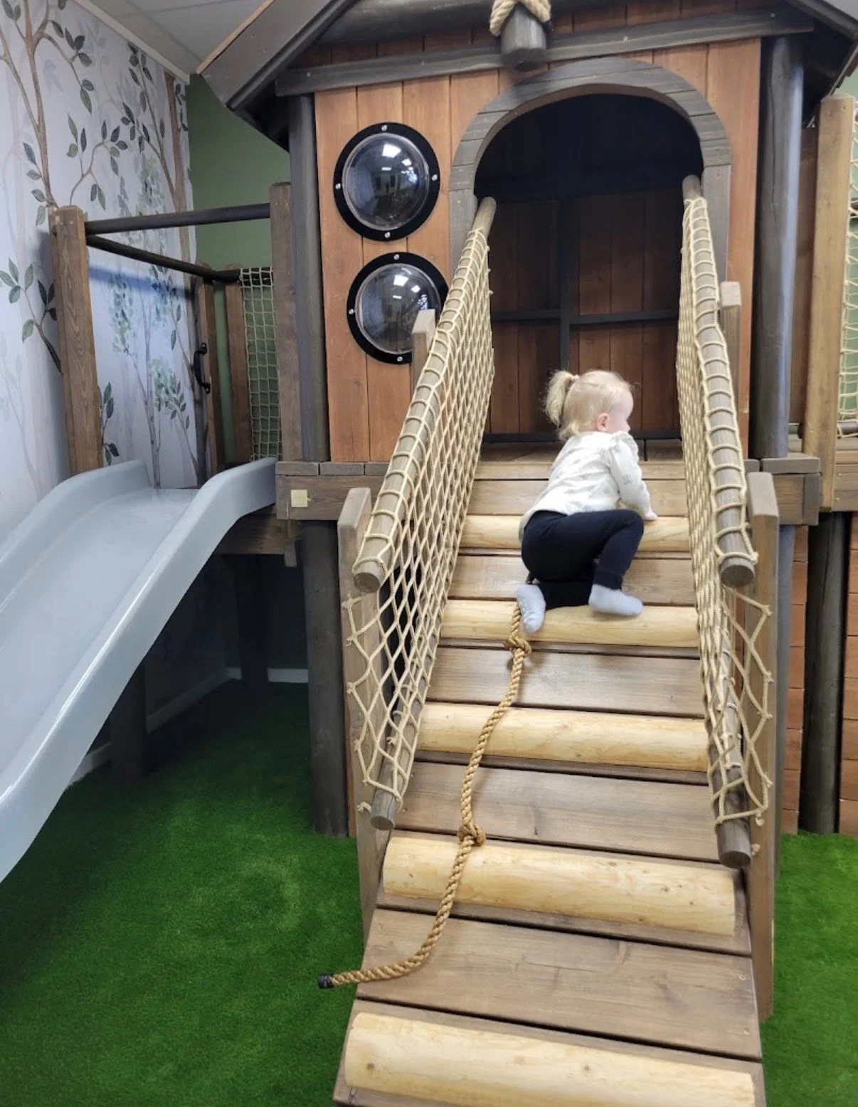Young child climbing a wooden playhouse with rope ladder, small slide on the side, and play windows, in an indoor play area with green artificial turf.