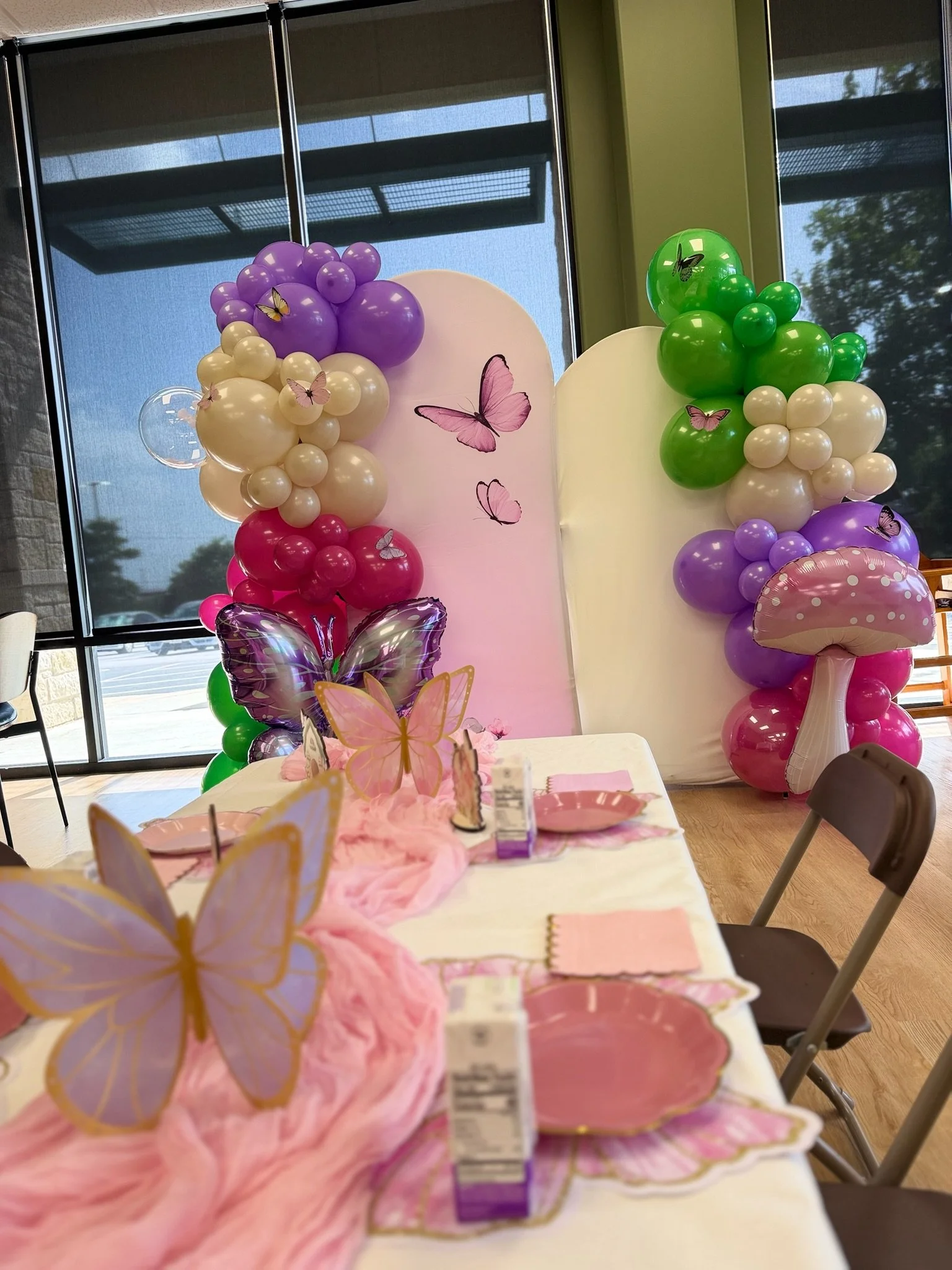 Decorative setup with pink, purple, green, and white balloons, some with butterfly stickers, behind a table with pink cloth and butterfly decorations, in front of large windows.