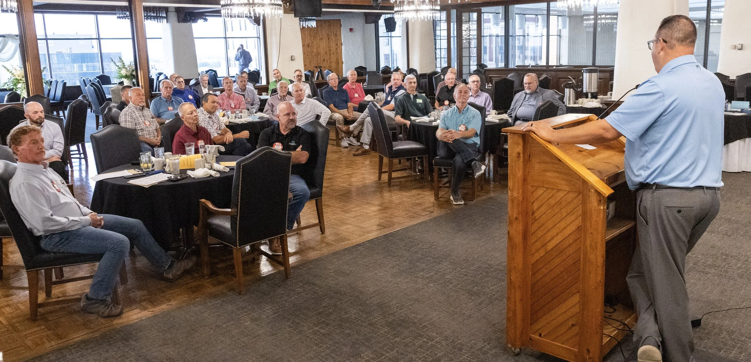 A presenter at a wooden podium addresses Wichita Executives’ Association members seated at round tables.