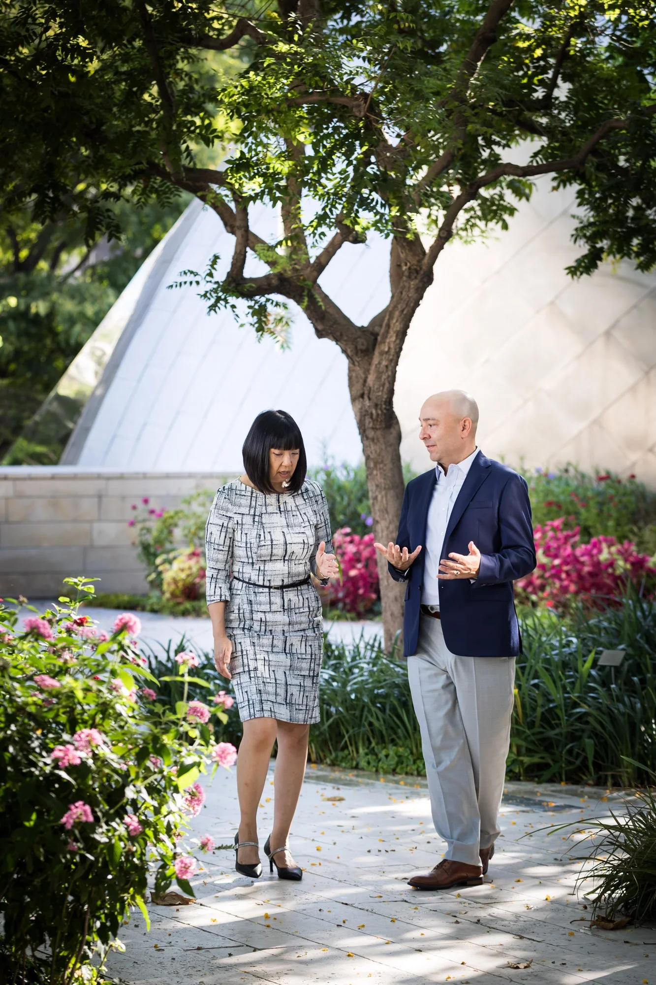 Career coach, Chris Lake, and a clientwalking and talking in a garden with pink and purple flowers, green bushes, and a large tree, with a modern building in the background.