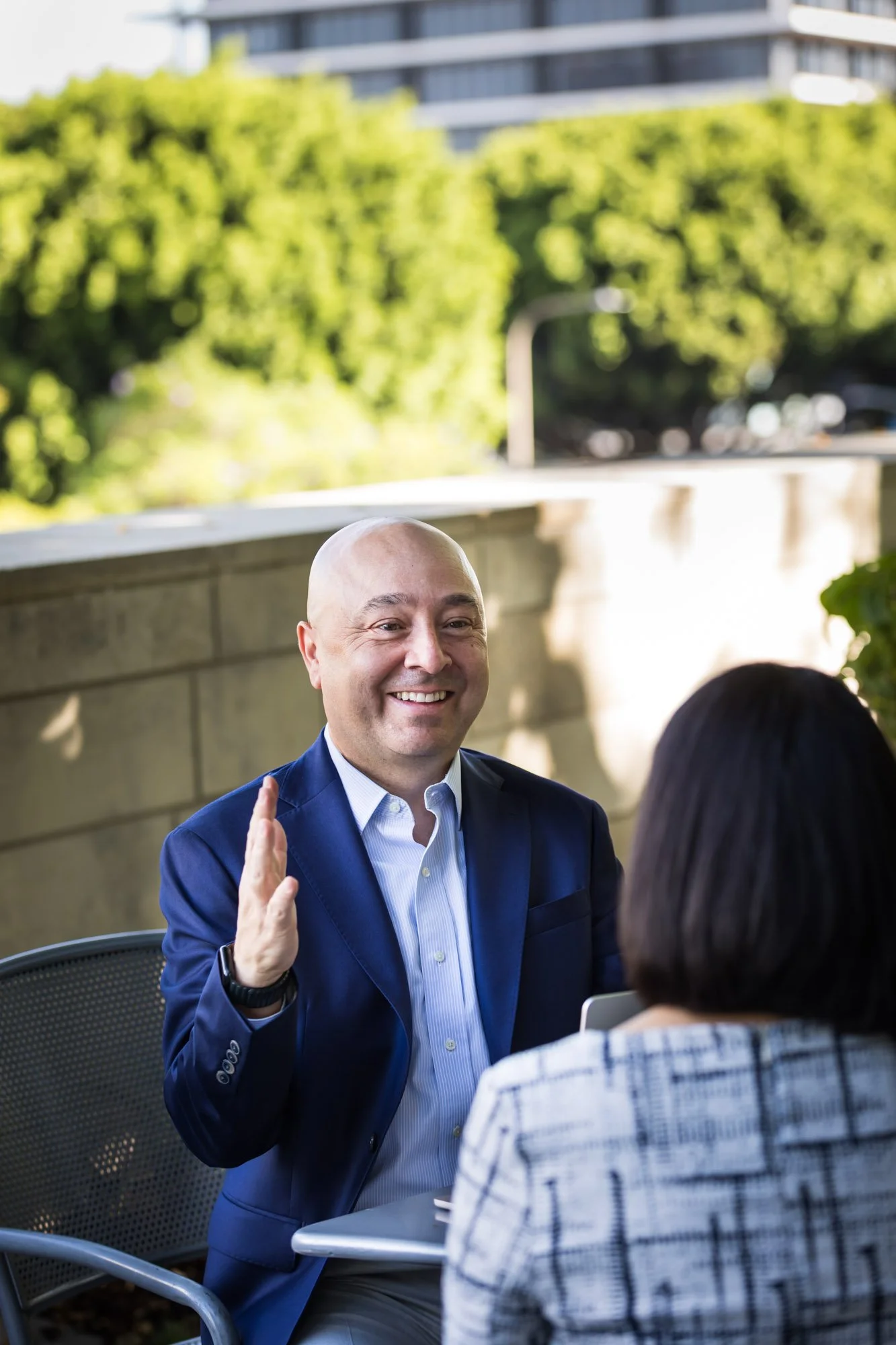A bald man wearing a blue suit, white shirt, and a smartwatch smiling and raising his hand while talking with a woman who has short black hair and is wearing a plaid shirt, seated at an outdoor table with a laptop. In the background, there is a green, leafy tree and a modern building.