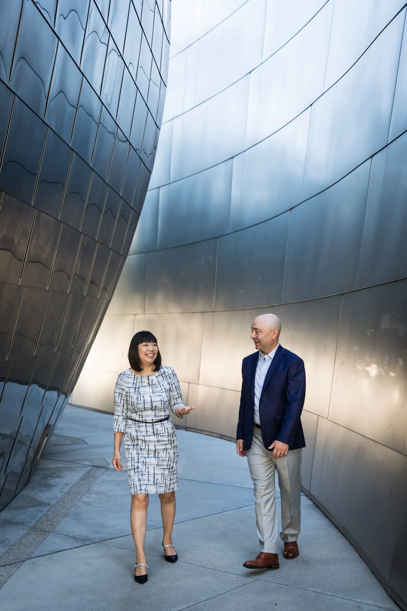 Two professionals, a woman and a man, walking and talking outside near a modern building with reflective metallic walls. The woman is smiling, wearing a patterned dress, and holding glasses in her right hand. The man is dressed in a blue blazer, white shirt, beige pants, and brown shoes, and is smiling at the woman.