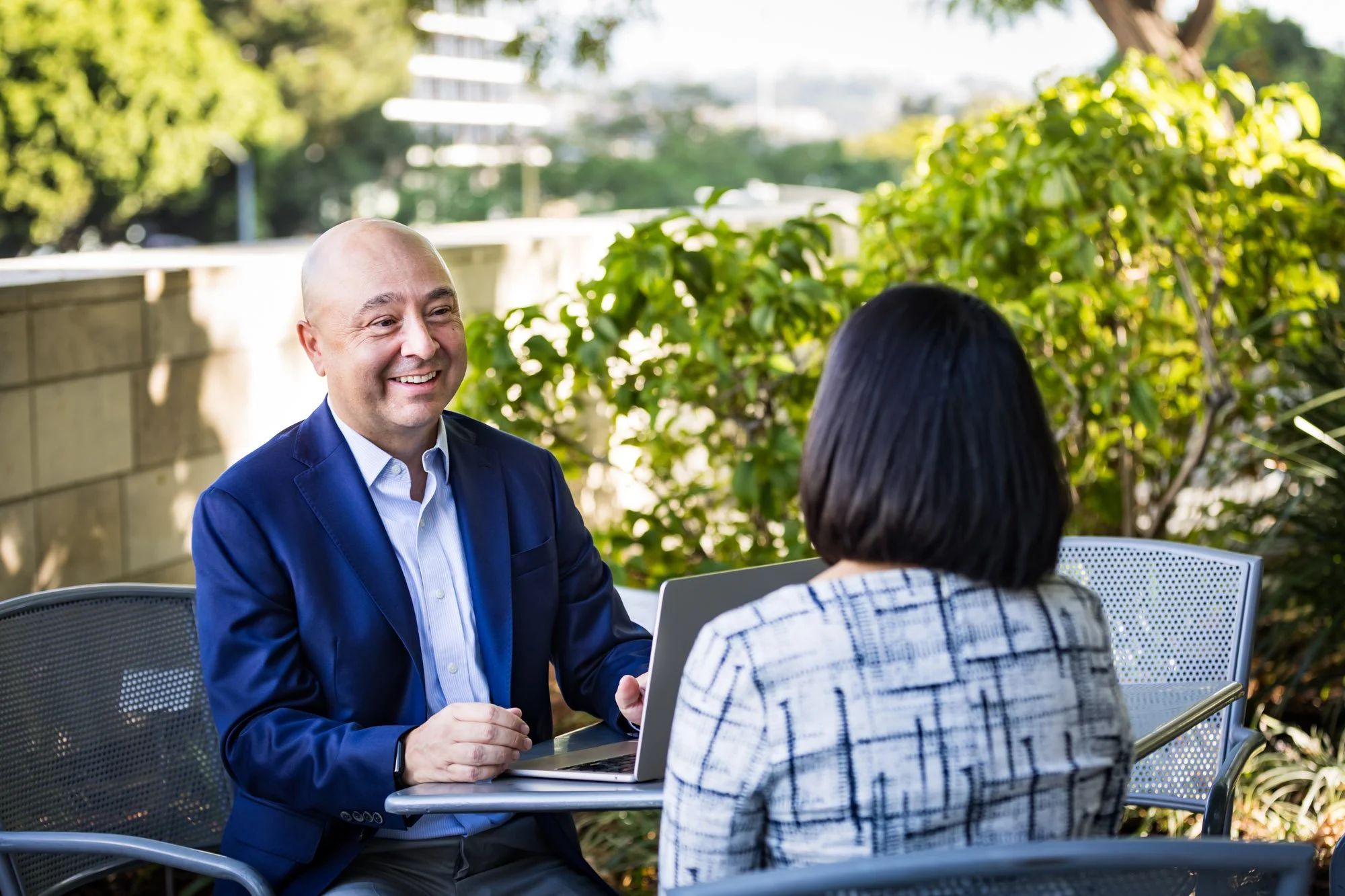 Career coach, Chris Lake, talking to a client with dark hair and a checked shirt, sitting outdoors at a table with a laptop, with greenery and trees in the background.