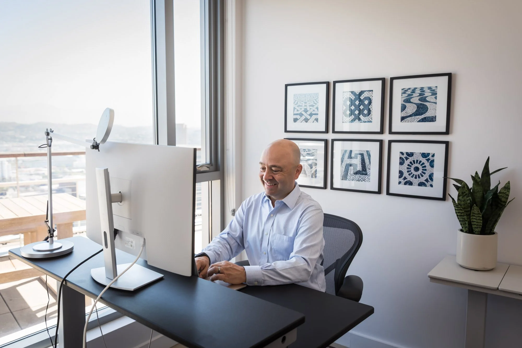 A man with a bald head sitting at a desk in front of a computer monitor in a modern office with large windows. There are six framed abstract art pieces on the white wall behind him and a potted plant on a white side table.