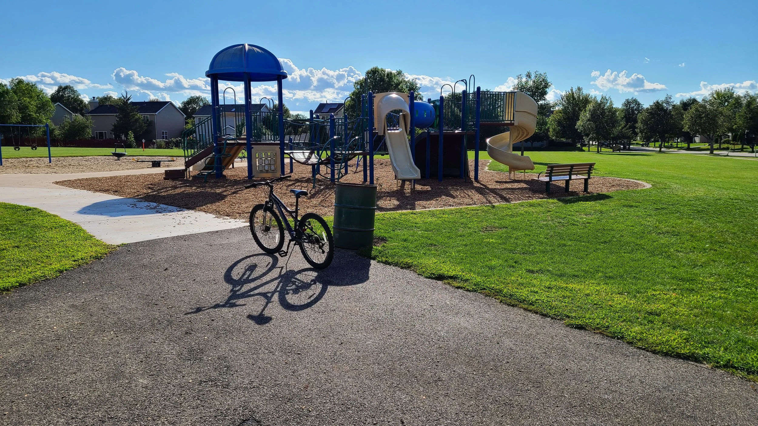 A playground with slides, swings, and climbing structures surrounded by green grass, trees, and residential houses in the background. A bicycle is parked near a trash can in the foreground. There is a pond a gazebo and half-court basketball trails