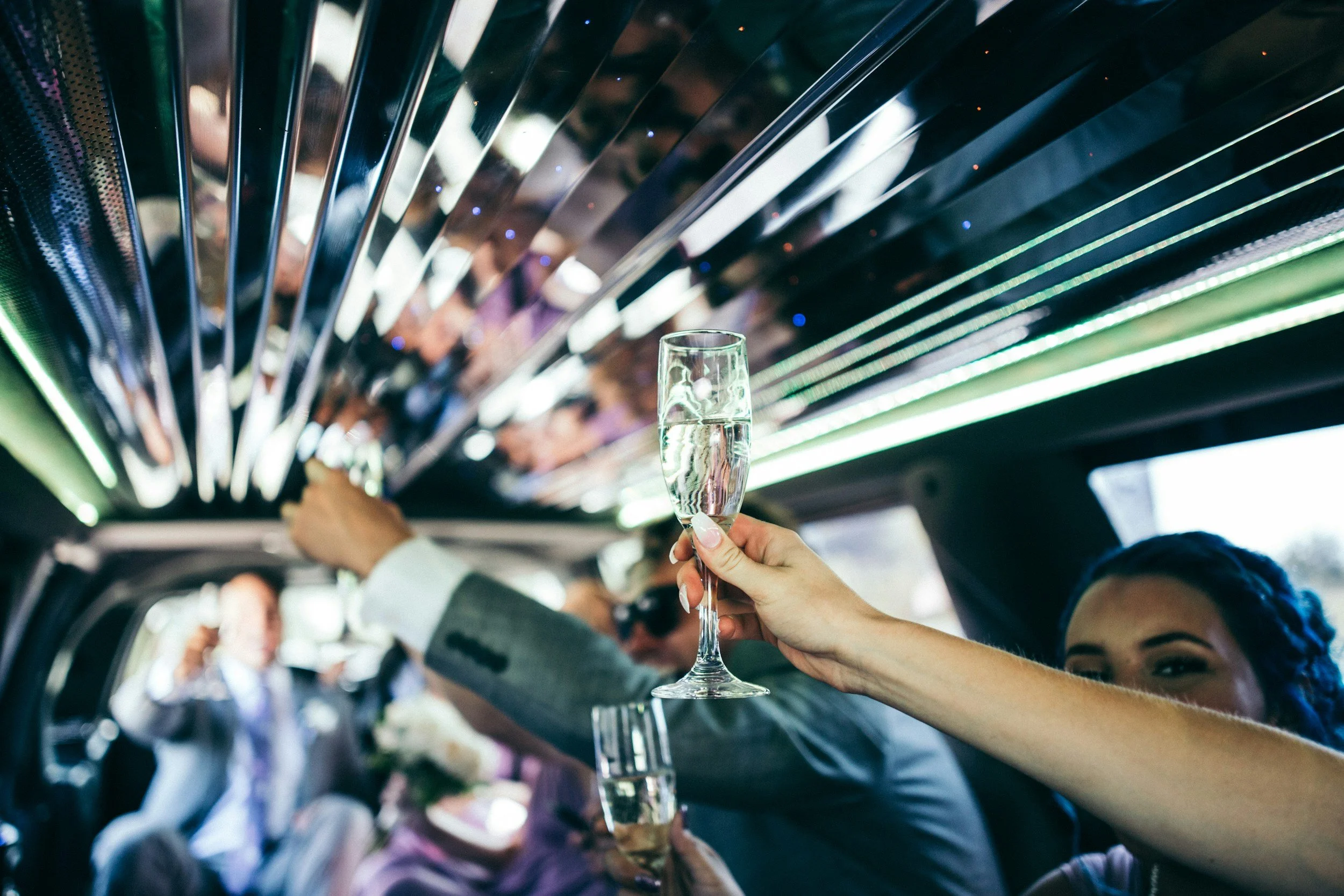 A person holding a champagne glass filled with a clear beverage inside a bus or train with other passengers. The ceiling has reflective, metallic, and illuminated striped design elements.