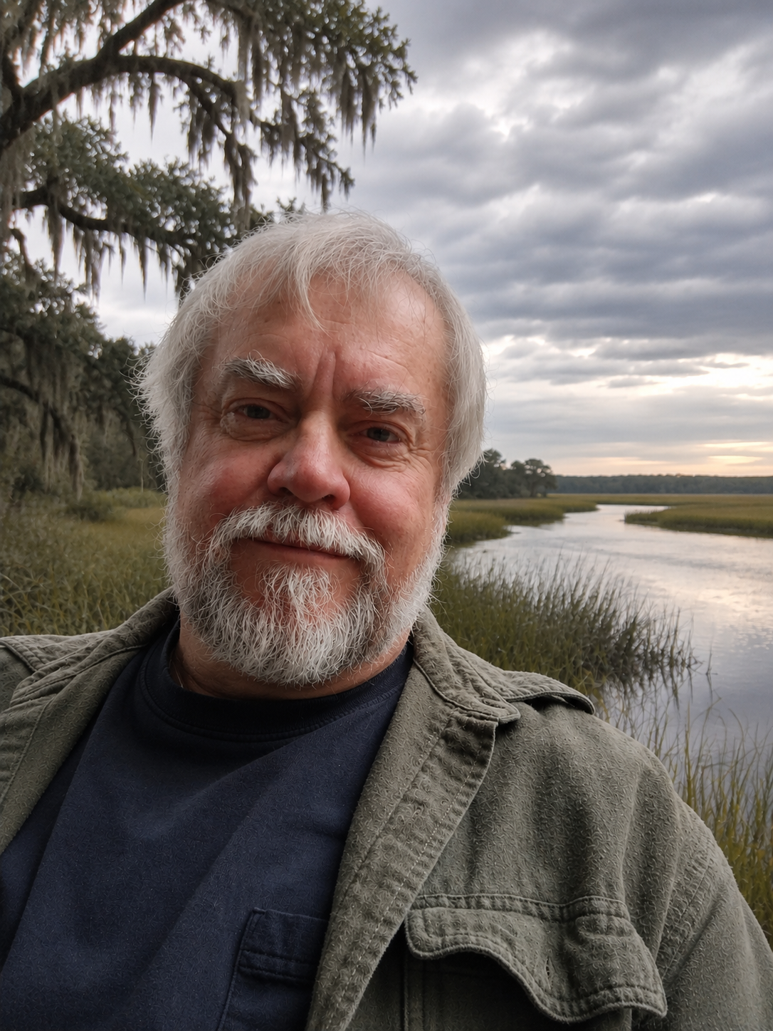 B Merrill. Jr. Author, a selfie outdoors near a marsh, with trees and overcast sky in the background.