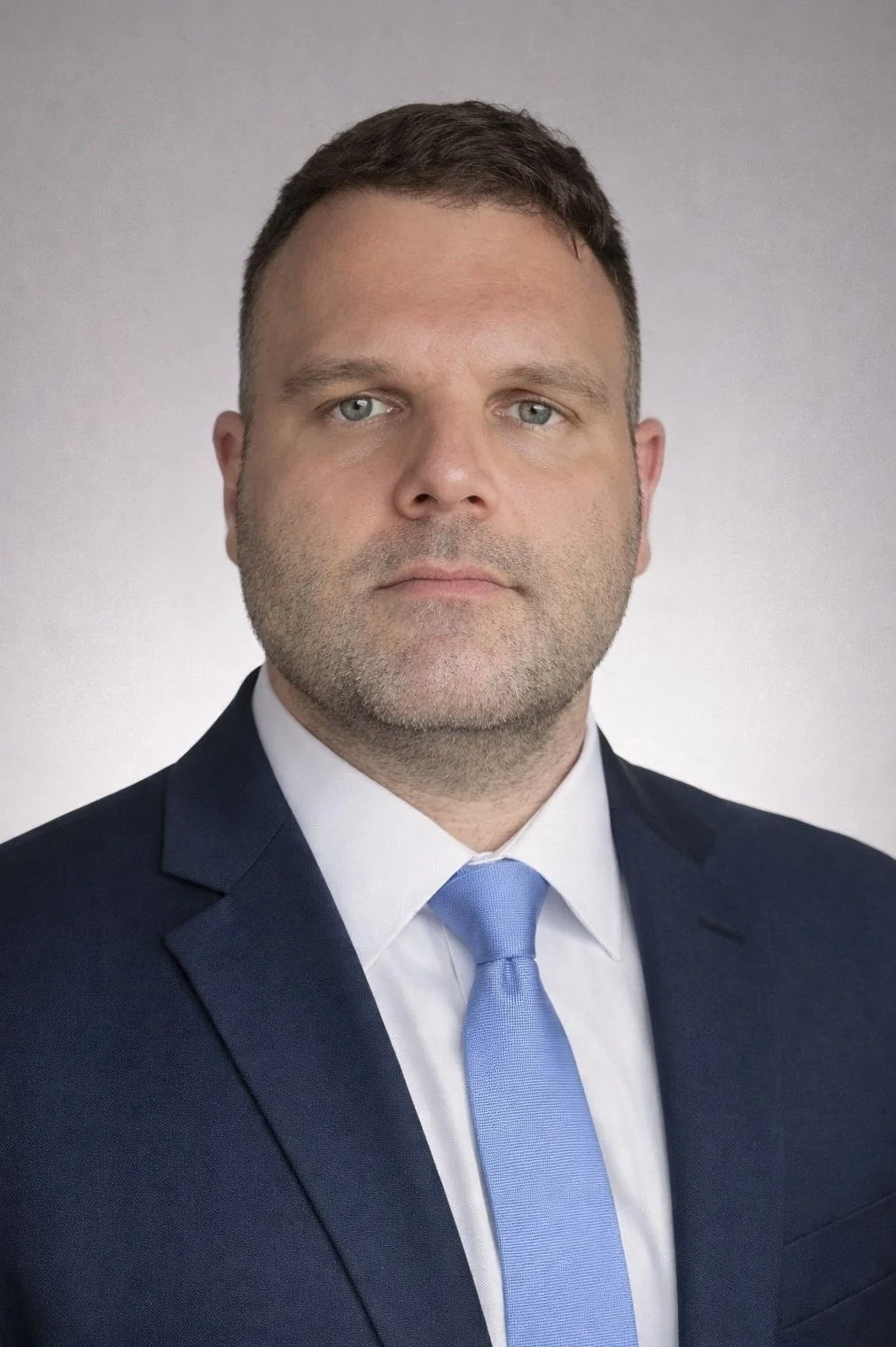 A professional headshot of a man in a dark suit, white shirt, and a light blue tie, posing against a plain gray background.
