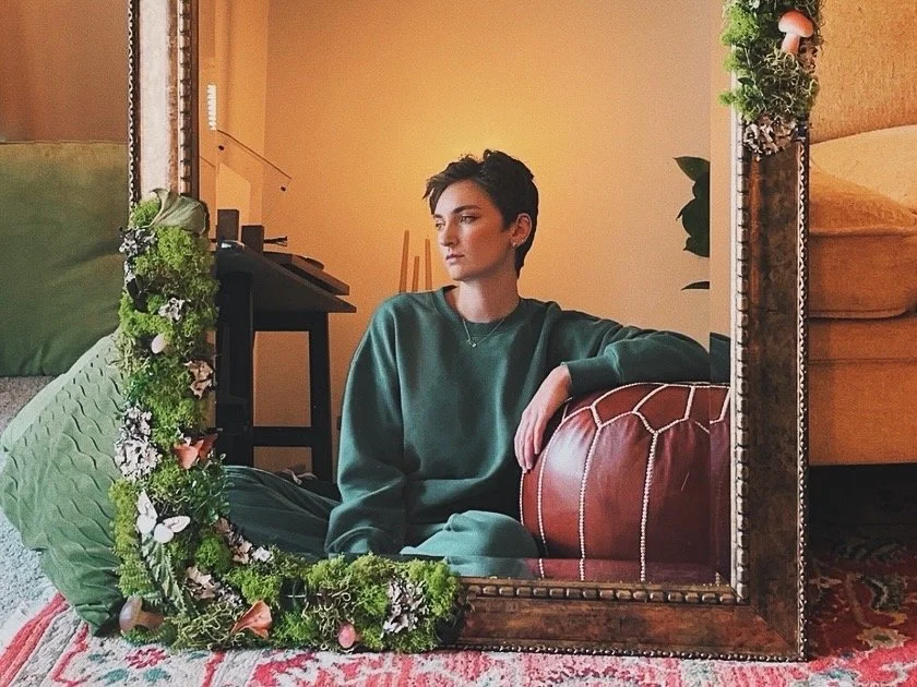 A woman sits on the floor, looking thoughtfully into a large mirror framed with greenery and flowers. She wears a dark green sweatshirt and has short dark hair, in a cozy, well-decorated room with a yellow wall, a couch, a patterned rug, and a table with shelves.