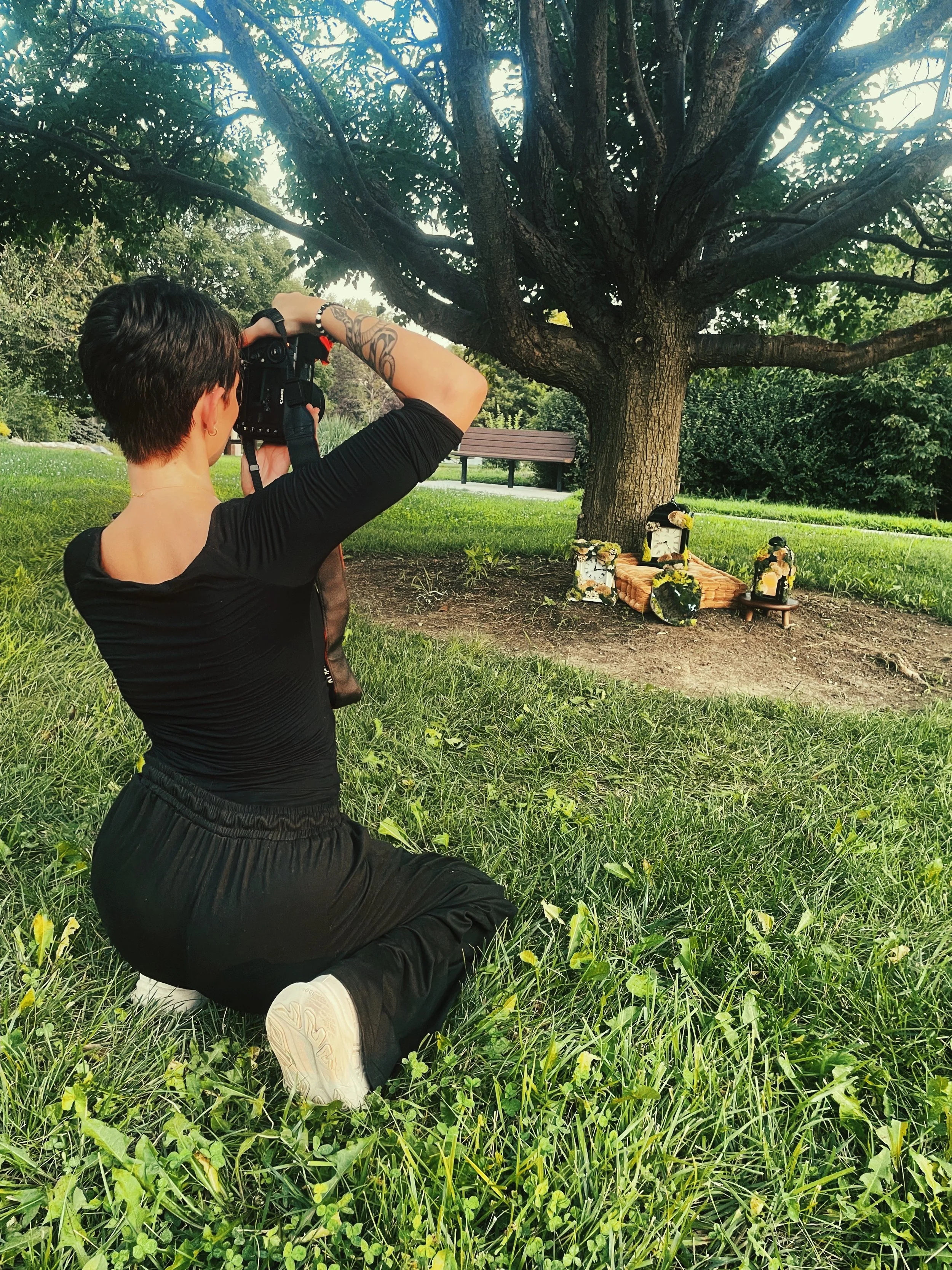 A person kneeling on the grass under a large tree, taking a photo or video with a camera. There are decorative items and photographs on a small table or platform near the base of the tree, suggesting a memorial or tribute area.