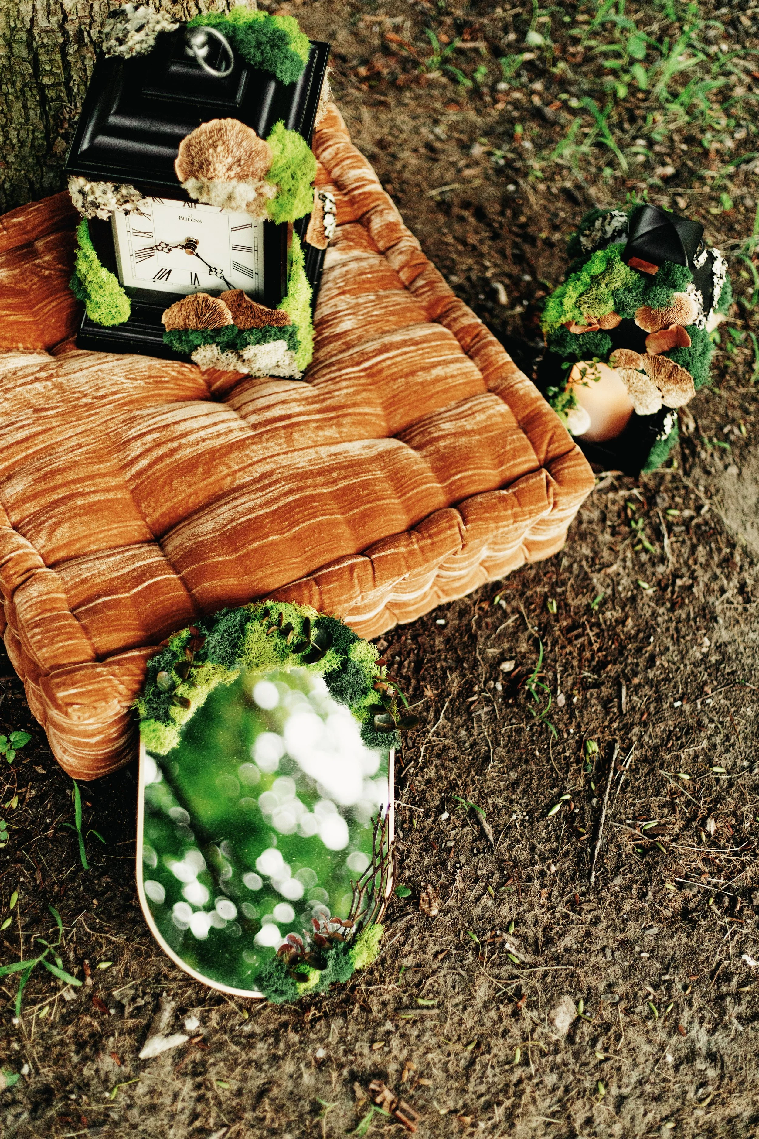 A clock with Roman numerals resting on a vintage orange velvet tufted sofa, decorated with green moss and fungi, situated outdoors on a patch of dirt and small plants.