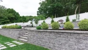 A landscaped backyard with a stone retaining wall, stairs, green grass, and various shrubs and bushes, enclosed by a white fence and surrounded by tall trees.