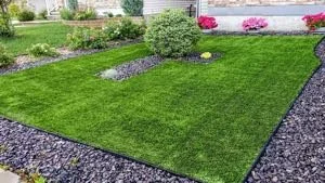 A well-maintained rectangular garden with lush green grass, surrounded by a border of black gravel, with flowering plants and shrubs in the background.
