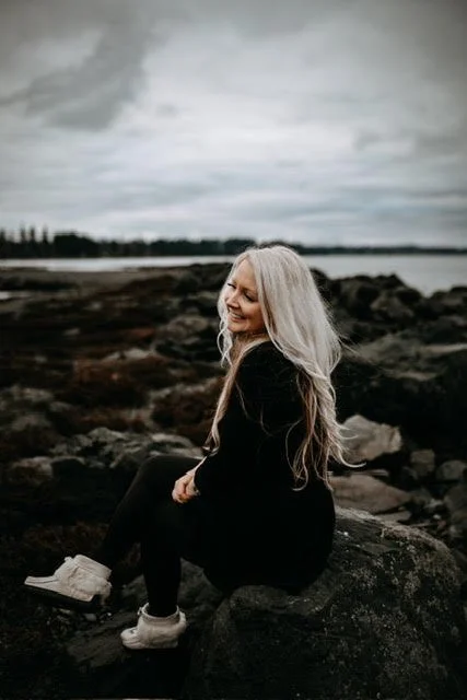 A woman with long gray hair sitting on rocks by a body of water under a cloudy sky