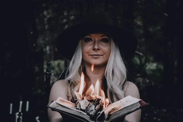 A woman wearing a black witch hat holding an open book with flames emerging from its pages, outdoors in a forest setting.