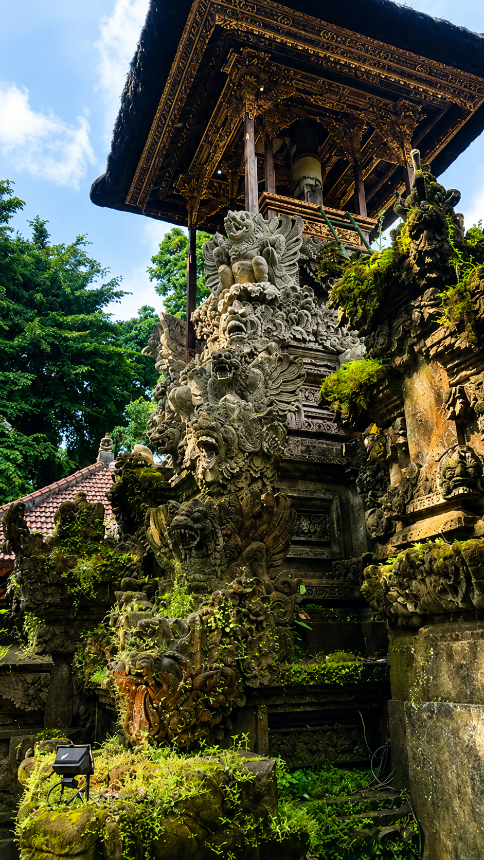Traditional Balinese temple with intricate stone carvings of mythical creatures and dragons, surrounded by greenery and moss, with a wooden structure and thatched roof at the top.
