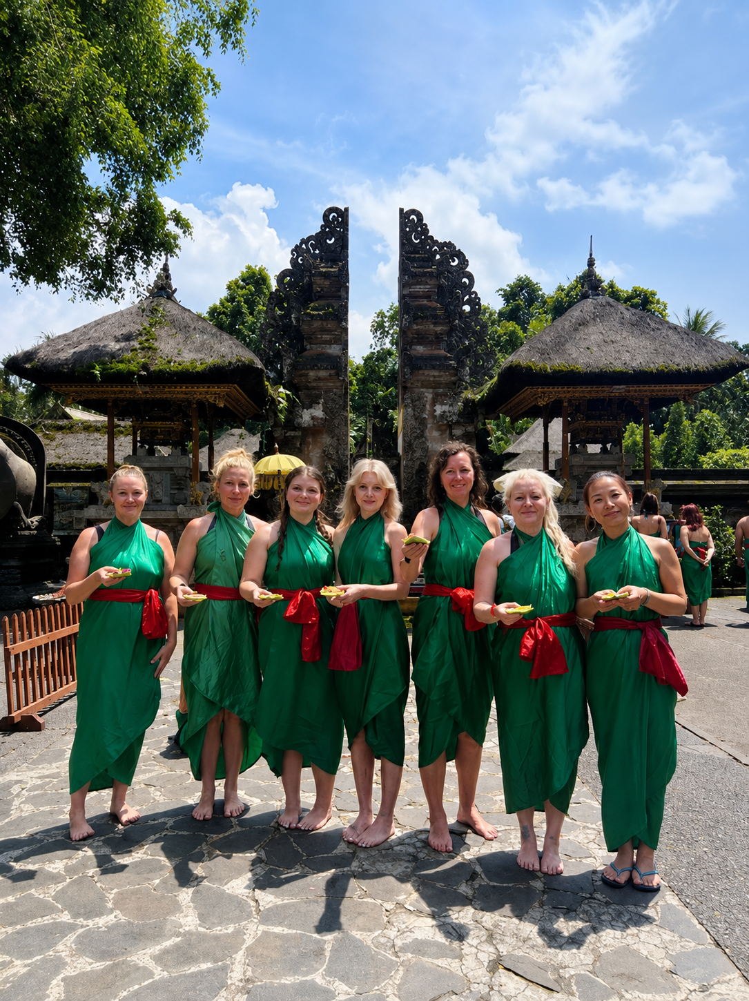 Group of women dressed in green traditional dresses with red sashes standing barefoot on stone pavement in front of a traditional Balinese entrance with thatched roofs and ornate carvings, holding small items in their hands during daytime.