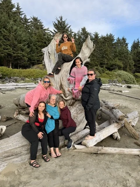 Group of six women posing around a large piece of driftwood on a beach, with trees in the background. One woman is sitting on top of the driftwood, making a peace sign. The others are standing or sitting around it.