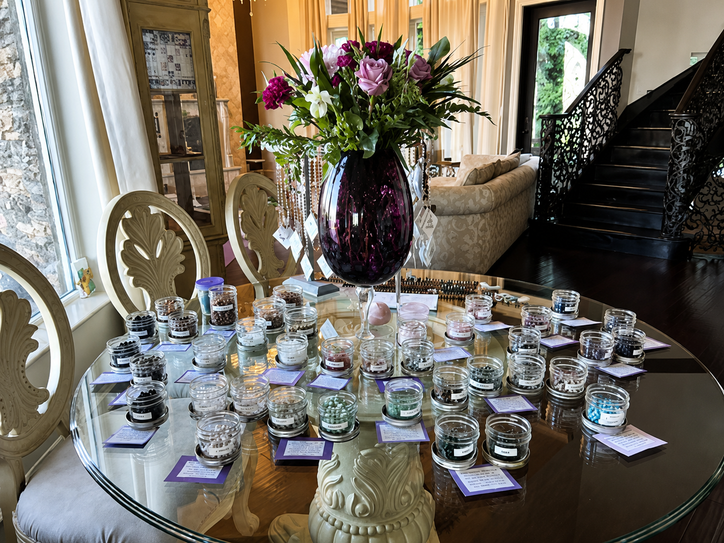 Table with a large purple flower arrangement, surrounded by small glass jars, each with different items, in a well-lit living room with a staircase and a sofa visible in the background.