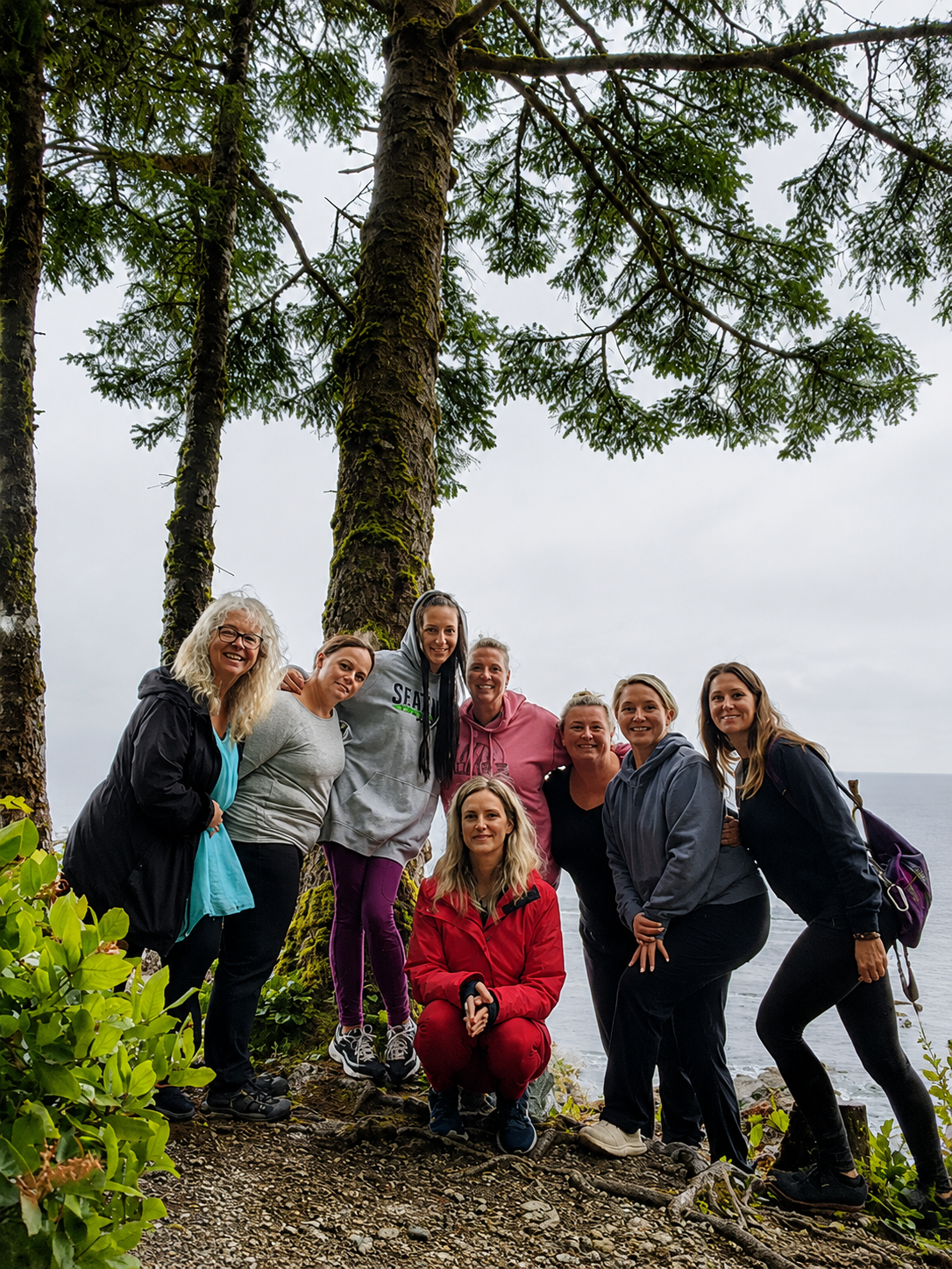 Group of nine women in outdoor clothing standing on a trail near tall trees with a body of water in the background.