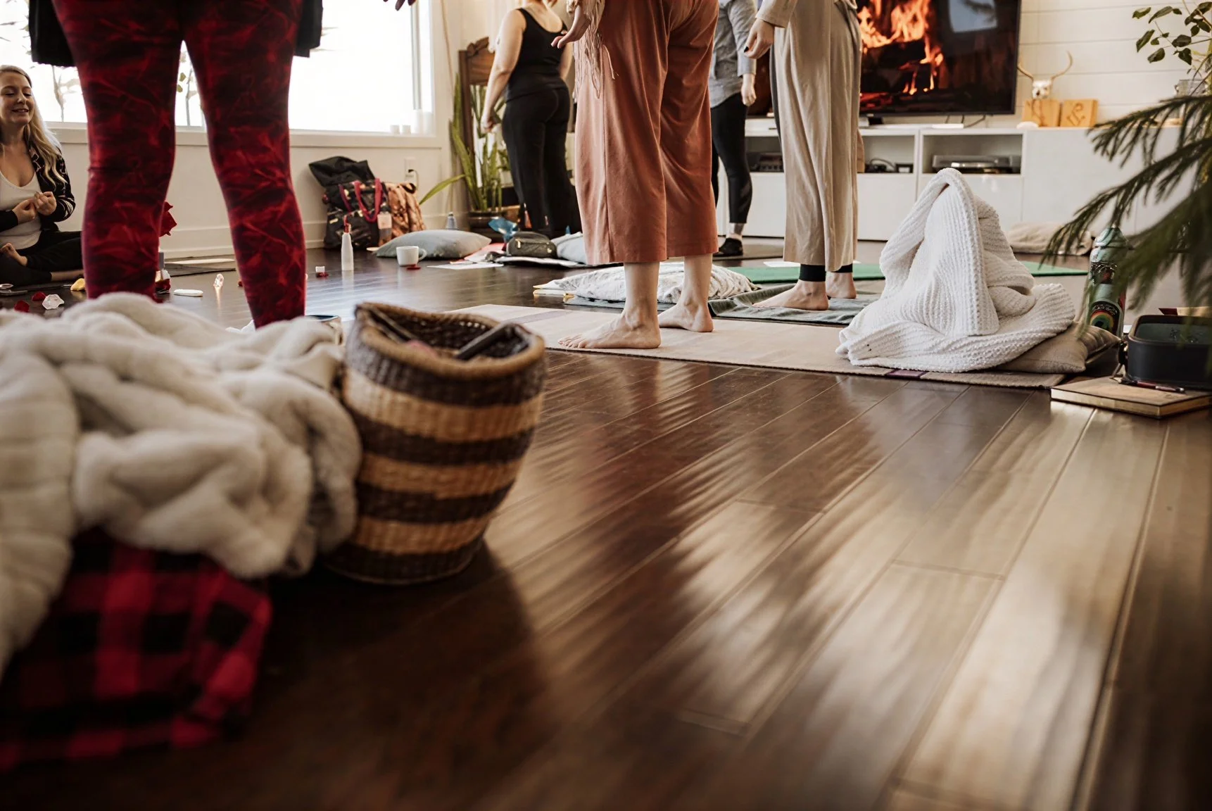 People participating in a yoga class in a cozy home setting, with some standing on yoga mats and a person sitting by a window.