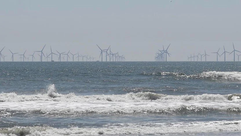 Ocean with wind turbines in the distance, waves crashing near the shore under a cloudy sky.