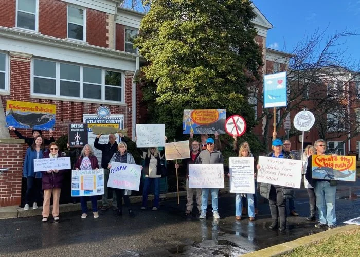 Group of people holding protest signs about climate change and environmental issues outside a brick building on a sunny day.