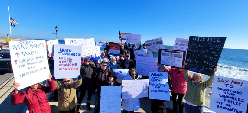 Group of people protesting on a pier holding signs against wind turbines, high taxes, and ocean pollution, near the ocean with a clear blue sky.