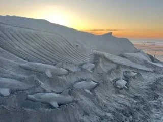 Snow and ice formations on a beach at sunrise or sunset with calm ocean waves in the background.