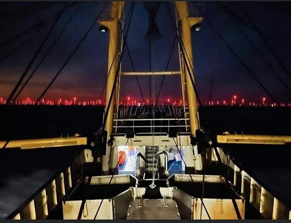 View from the deck of a ship at night showing the ship's structure, rigging, and a city skyline with red lights in the distance.