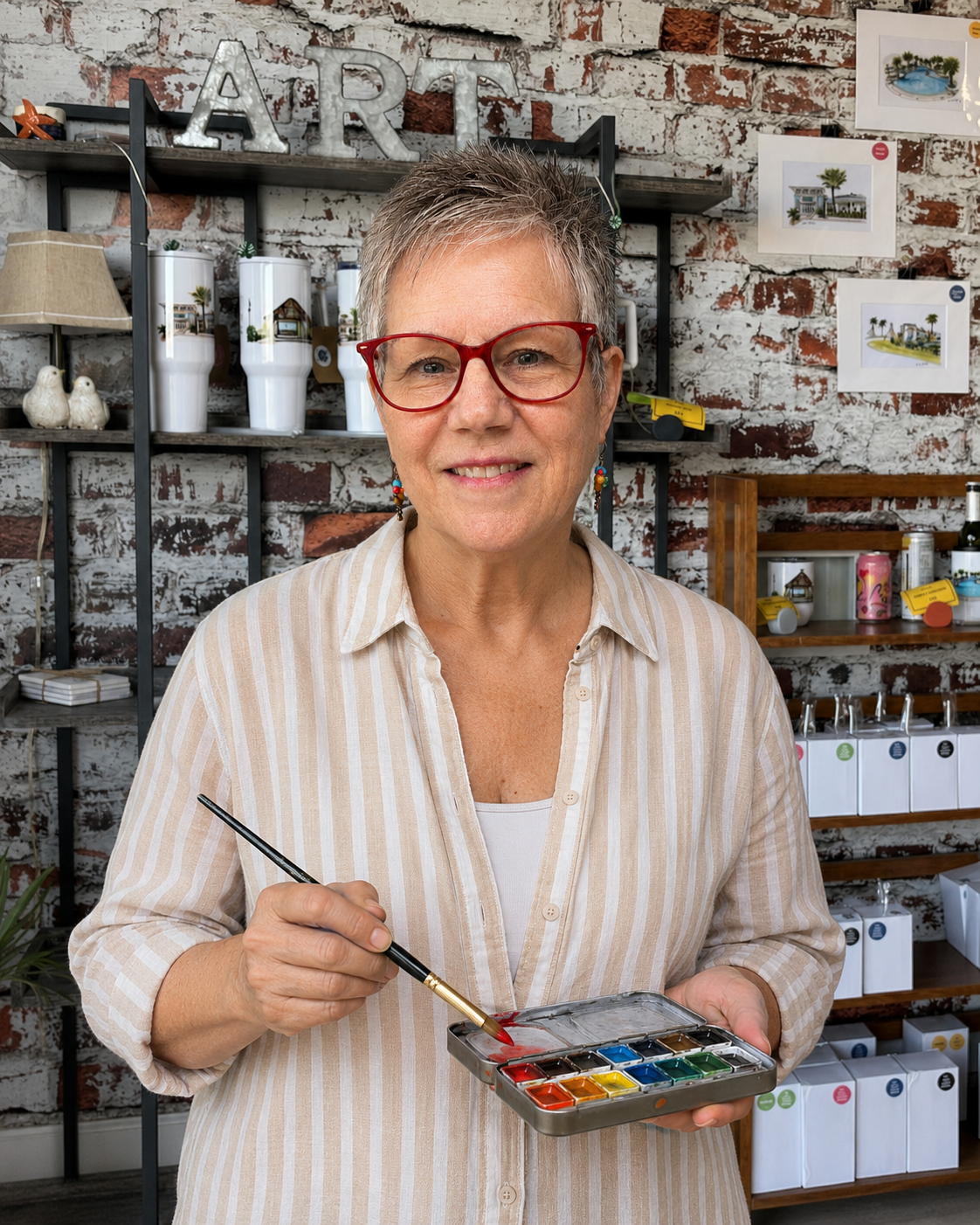 A woman with short gray hair and red glasses painting with watercolors in an art studio with a brick wall background.