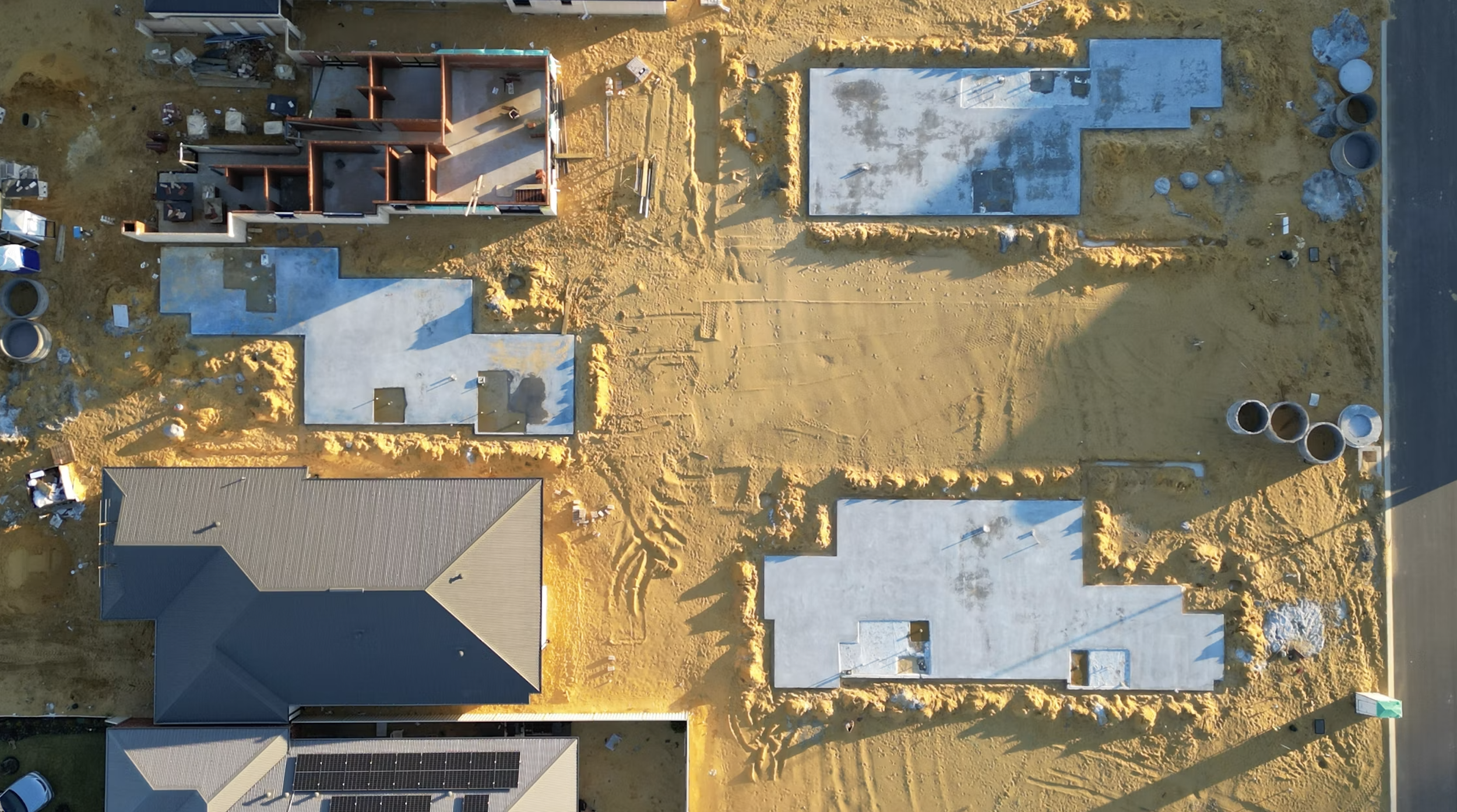 Aerial view of a construction site showing parts of multiple houses, with the presence of dirt, construction materials, and equipment, as well as the ongoing framing and foundation work.