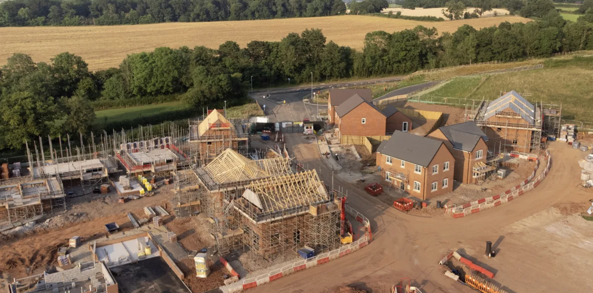 Aerial view of a construction site with multiple houses being built, scaffolding and wooden frameworks, surrounded by trees and fields.