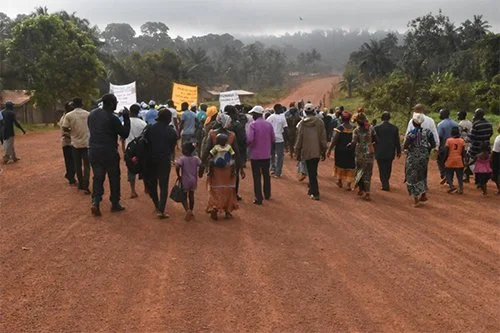 A group of people marching on a dirt road holding signs, with greenery and trees on both sides, under cloudy skies.