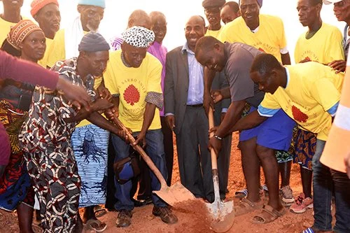 Group of people, including women and men, participating in a groundbreaking ceremony, some wearing yellow shirts with a flower logo, others in traditional clothing, holding shovels to dig into the dirt.