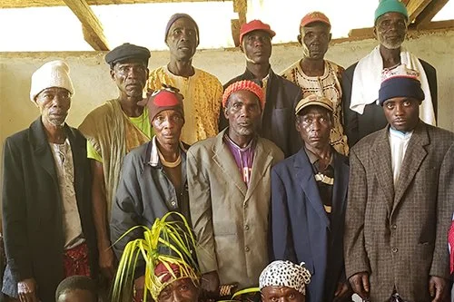 Group of eleven men and women standing indoors, some wearing hats and traditional clothing, with a plant in the foreground.