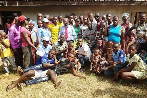 A large group of people, including men, women, and children, gathered outdoors on grass in front of a building.