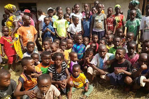 A large group of children and a few adults gathered outdoors, some sitting on the grass and others standing, in front of a building with a plain wall.