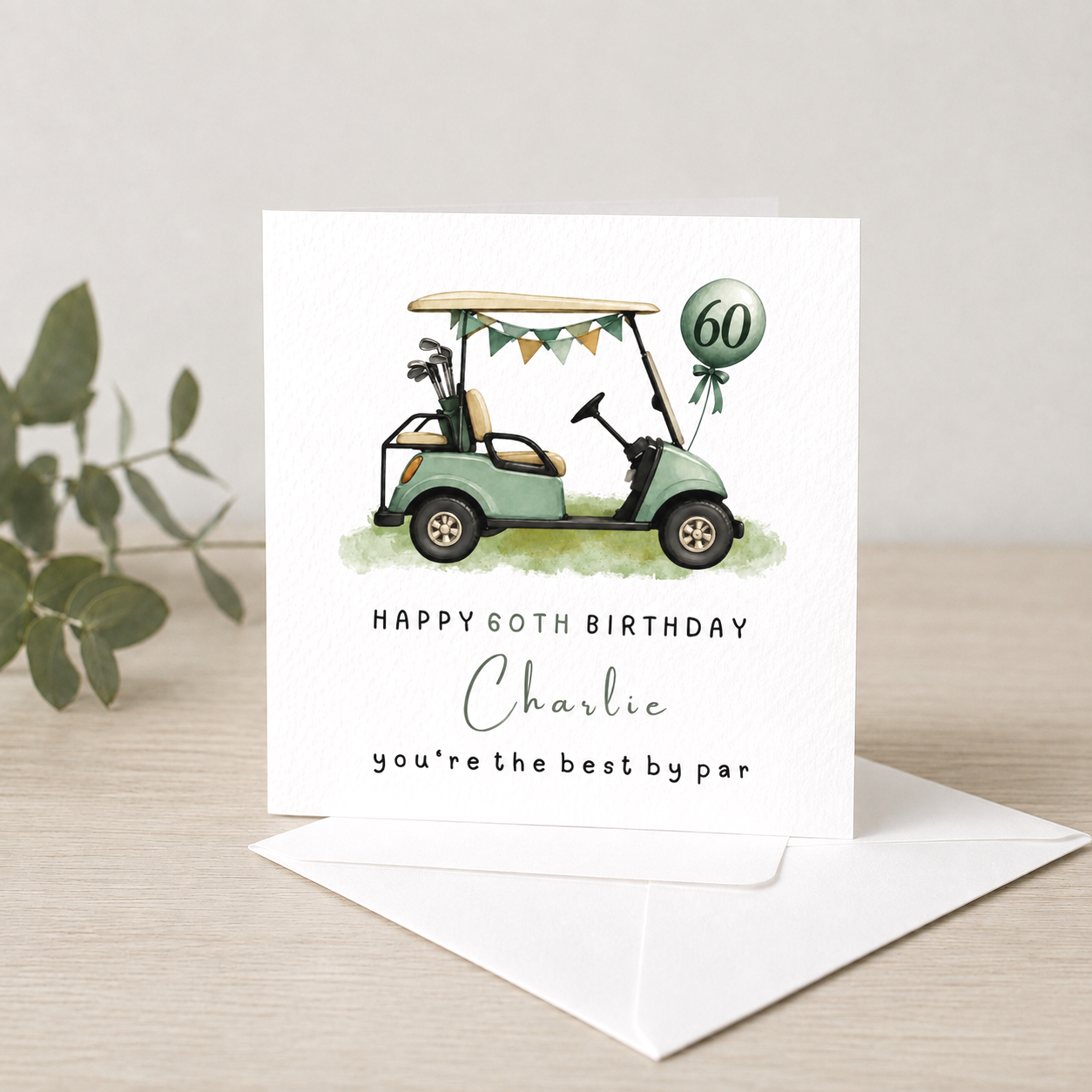 A birthday card with a golf cart illustration decorated with bunting and a balloon marked 60, sitting on a light wood surface with a leafy branch nearby.